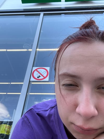 A close-up, slightly angled, indoor shot depicts a person with reddish-brown hair pulled back in a bun, wearing a purple shirt. Positioned behind the person and partially visible is a window with a "No Smoking" sign affixed to it. Based on the window's reflection, the exterior appears to be a bright, possibly overcast day. The specific location in Казань, Russia cannot be determined from this image.