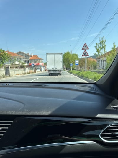 View from inside a vehicle, presumably a car, looking out the windshield. The vehicle is traveling on a road in what appears to be a residential or commercial area. A large white truck is visible in front, along with buildings on either side of the road. Road signs indicating pedestrian crossings and traffic conditions are present. The sky is clear and blue. The interior of the vehicle includes a dashboard with air vents and textured surfaces. The location is Daskotna, Bulgaria.