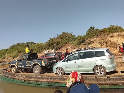 This image depicts a ferry operation on a body of water in Ibi, Nigeria. The scene is observed from the ferry deck, which features wooden planks and a green hull visible at the water line. Two primary vehicles occupy the ferry: a dark blue Toyota Hilux pickup truck, displaying "TOYOTA HILUX" on its side, and a light blue/silver minivan positioned to its right. A person wearing a yellow shirt and green shorts is seated on the roof rack of the Toyota Hilux. Behind the Hilux, a red vehicle is partially visible. The immediate foreground shows the back of a person's head and shoulder, wearing a multi-colored floral headscarf (red, white, yellow) over a blue garment. To the right, a sandy, vegetated riverbank slopes upwards, populated by several individuals, some wearing red clothing, suggesting activity related to the ferry's landing or departure. The sky above is clear and light blue.