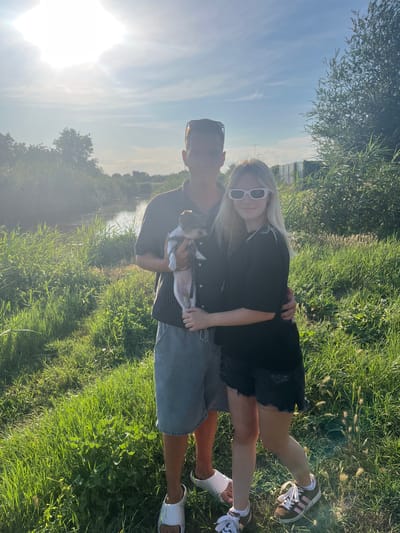 A young couple, a male and a female, are standing in a grassy field near a body of water, possibly a canal or river. The male, on the left, is wearing a striped short-sleeved shirt, grey shorts, and white slides. He is holding a small dog in his arms. The female, on the right, is wearing a black t-shirt and black denim shorts. She has blonde hair and is wearing white sunglasses. The sun is shining brightly, casting a warm glow over the scene. The background consists of lush green grass, trees, and the water. The overall impression is a casual, outdoor moment. The location is identified as Yanovo-Grushevskiy, Russia.