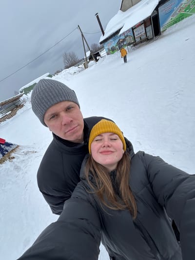 A selfie image depicts two individuals in a snowy outdoor environment identified as the Zaosinovo Ostrich Farm in Zaosinovo, Vologda Oblast, Russia. In the foreground, a male subject wears a grey knit beanie and a black puffer jacket, looking directly at the camera with a neutral expression. Positioned in front of him and to the right, a female subject wears a yellow knit beanie and a black puffer jacket; her eyes are closed, and she displays a slight smile. The ground is uniformly covered in deep snow under an overcast grey sky. In the mid-ground, a wooden utility pole with overhead lines is visible. To the right, a building with a snow-laden roof features multiple signs in Russian, prominently displaying "СТРАУСИНАЯ ФЕРМА" (Ostrich Farm), "ЭКСКУРСИИ" (Excursions), and a phone number (278 15 36). A small individual in a bright yellow jacket is walking away from the camera, further down a snowy slope toward this building. A partially obscured blue and white patterned object rests on the snow near some dry vegetation on the far left.