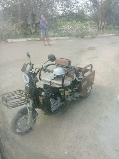 In the foreground of the image is a black three-wheeled electric scooter. It has a front basket, a single headlight with a white ring, and two seats. A white helmet sits on the passenger seat. The scooter is parked on a gravel surface.

In the background, a man stands with his hand on his chin, appearing to be deep in thought. He is wearing a blue t-shirt and grey shorts. He is barefoot, wearing flip-flops. Behind the man, there are trees and vegetation, with some debris scattered on the ground. The scene appears to be outdoors in a somewhat arid environment. The lighting suggests it could be daytime, possibly overcast. The overall impression is casual and somewhat still.

The scooter has "Neo hlike" visible in white text on its side.