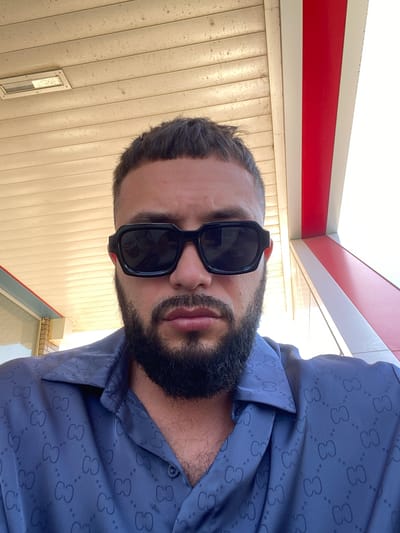 A man with short dark hair, a beard, and sunglasses is pictured from the chest up. He is wearing a blue button-down shirt with a repeating dark pattern. The background features a beige, corrugated ceiling with a light fixture. To the right, a red and white striped structure is visible, suggesting an outdoor setting. The image appears to have been taken at a gas station in Korkmaskala, Russia.
