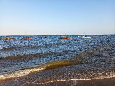 The image shows a calm ocean scene taken from a beach near the shore, with several small sailboats anchored in the water. The beach is sandy and light brown, with a gentle wave breaking on the shore. The water is blue and calm, with small ripples and waves. There are no people in the scene. The photo was taken at a location in the city of Maputo, Mozambique, with the coordinates of -25.952 latitude and 32.614 longitude.