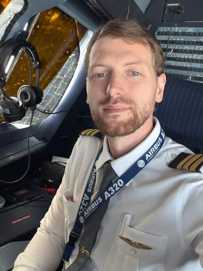 A pilot wearing an Airbus A320 lanyard and pilot epaulets is seated in the cockpit of an aircraft. The cockpit features an array of control panels and a headset, indicating the pilot is in a work environment. The exterior visibility from the cockpit suggests the plane is near the ground, possibly during taxi or parked on the tarmac. Labels for "oxygen mask" and "ADIRS" are visible on panels within the cockpit. Without additional environmental identifiers or context, pinpointing a specific location such as Schiphol is not definitively possible.