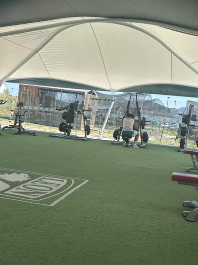 An outdoor gym features various weight machines on a green turf floor. A person uses a seated chest press. Surrounding the gym is a fence and netting. Part of a white tent structure is visible in the foreground. The gym appears to be located on school grounds with the school logo painted on the turf.