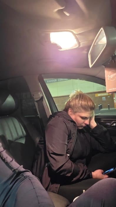 The image captures the interior of a vehicle at night, positioned outside a retail establishment identified as a "GARDEN CENTRE" in Newry, United Kingdom, specifically likely the B&Q store.
A single adult female is seated in the front passenger seat. She has light brown hair styled upwards and is wearing a dark, possibly black, padded jacket over a dark top. Her head is tilted downwards and to her right, with her right hand resting near her face. She is actively viewing a smartphone held in her left hand, which emits a blue light from its screen.
The car's interior dome light is illuminated, casting light on the ceiling and passenger area. A rearview mirror is visible, from which a brown-colored air freshener labeled "FRAGRANCE" and "ONE MILLION" hangs. In the lower left foreground, the legs of a second individual, wearing blue trousers, are partially visible, suggesting another occupant within the vehicle.
Through the passenger side window, the exterior environment reveals part of a large, light-colored building with horizontal paneling. Green lettering on the building clearly displays "GARDEN CENTRE", with an "ENTRANCE" sign visible directly below. The exterior appears dimly lit, consistent with nighttime conditions.