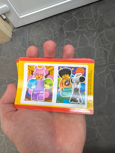 A hand holds a stack of three colorful cards with Lego-style characters. The top card features a character with a pink wig and fluffy ears, holding a blue creature and a green creature. The second card depicts a character in an orange shirt with a rocket design, standing next to a telescope. The third card is partially visible. The cards are set against a backdrop of grey tiled flooring and a white door frame, suggesting an outdoor or semi-outdoor setting, possibly near a building entrance. The image was taken at the entrance of a building in Suceava, Romania.