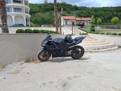 The image shows a black Yamaha motorcycle parked on a paved surface. A black helmet sits on the seat of the motorcycle. The background includes a beige wall and a set of wide stairs leading up to a landscaped area with green grass, trees, and several houses with red tile roofs. The location is Pisanets, Bulgaria.
