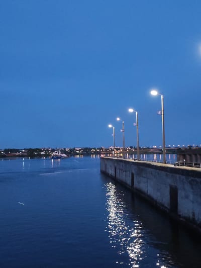 The image depicts a waterfront scene at dusk in Sheksna, Russia. A concrete embankment with several lampposts lines the right side of the frame, extending into the distance. The lampposts are lit, casting reflections on the dark, still water. In the distance, across the water, a city skyline is visible with scattered lights. A boat, possibly a ferry or barge, is navigating the water, also illuminated by lights. The sky is a deep blue, indicating twilight or early night. The overall impression is one of a peaceful, urban waterfront setting during the evening.
