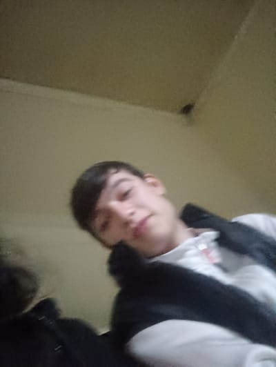 A selfie-style image depicts a young male with dark hair and light skin wearing a white shirt and dark jacket. He gazes slightly downward toward the camera. The background is a plain, light-colored wall, with an indistinct object visible in the upper right corner. Part of another person's dark clothing is visible on the left side of the frame. The image is slightly blurry and the perspective is from a low angle.