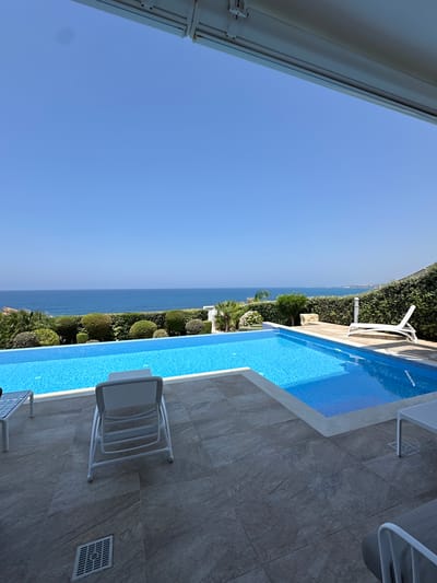 This is a photograph of a swimming pool area with a sea view, likely from a hotel or private residence. The pool is rectangular and filled with bright blue water. On the tiled patio area around the pool, there are several white lounge chairs, some folded and others set up for use. A white sun lounger is visible near the right side of the pool, on a raised wooden deck. A well-maintained hedge borders the pool area, separating it from the sea beyond. The sea is calm and extends to the horizon under a clear blue sky. Lush green vegetation, including trees and bushes, is visible along the coastline. The architectural element visible at the top of the image suggests a covered patio or balcony. The location is identified as Kissonerga Community, Cyprus.