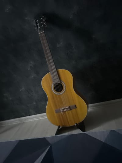 An acoustic guitar rests on a black stand against a dark, textured wall in what appears to be an indoor setting. The guitar is positioned at an angle, leaning towards the right side of the frame. Its body is a warm, light brown wood with visible grain. The soundhole is circular and surrounded by an ornate, light-colored rosette. The bridge is also visible, holding the strings in place. The neck of the guitar extends upwards, and the headstock, with its tuning pegs, is at the top left of the image. The floor in front of the guitar is a light-colored wood. The background wall has a slightly uneven, dark texture, creating a dramatic contrast with the guitar. The lighting appears to be dim, with shadows cast by the guitar. The lower portion of the image shows a patterned surface, possibly a piece of furniture or wall decoration, with geometric shapes in shades of grey and dark blue. No people are present in the image, and no specific location in Apsheronsk, Russia can be identified from the visual information provided.