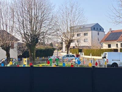 The image depicts a children's playground and residential street scene in Aumetz, France, observed on a clear, sunny day. In the immediate foreground, a dark gray or black solid fence stretches horizontally across the bottom of the frame. Behind this, a metal fence with vertical bars and colorful, stylized cutouts (red, blue, yellow, green) encloses a playground area.

Within the playground, from left to right, a green and yellow children's slide or climbing structure is visible. One adult female, wearing a light-colored top, is seated or crouching near this structure. Further right, a small green-roofed playhouse with white walls and a red door is present, alongside other brightly colored playground elements such as yellow and blue spring riders. The playground ground cover appears to be light-colored, possibly wood chips or sand.

Beyond the playground, a paved street runs horizontally across the midground. A silver minivan or estate car is visible on the left side of the street, and a white commercial van is parked on the right, partially obscured by a hedge and the playground fence.

The background features several residential buildings and mature, bare-branched deciduous trees, indicating a late winter or early spring season. To the left, a portion of a house with a reddish roof is seen. In the center-right, a multi-story white building with a distinctive gray metal gabled roof and multiple windows is prominent. To the far right, a house with a reddish-brown tiled roof is visible, notable for the several dark solar panels installed on its roof. Well-maintained green hedges line the properties along the street. A streetlamp with a yellow light fixture stands further down the street on the right. The sky is clear and light blue.