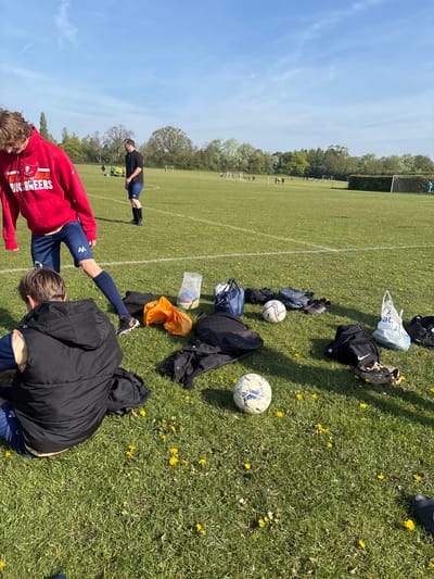 The image shows a grassy field with yellow dandelions, likely a sports field. There are soccer balls and bags scattered on the grass. In the foreground, two people are present: one wearing a red hoodie with "BUCCANEERS" printed on the back is stretching their leg, while another person in a black jacket is bending down. In the background, another person is stretching. Beyond them, a soccer game appears to be in progress in the distance. A white line marks the field and there are trees in the distance. The sky is blue with light clouds. The location is likely St Albans, United Kingdom.