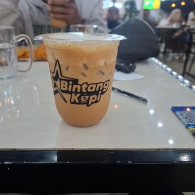 This media file captures a close-up, angled view of a takeaway coffee cup filled with a creamy, iced beverage. The cup is made of clear plastic and sealed with a plastic lid, some of which appears slightly peeled back. Inside, the drink is a light brown color with visible ice cubes and a frothy layer at the top.

The cup bears a black logo with stylized white lettering that reads "Bintang Kopi." The logo also features a star graphic. The cup is resting on a light-colored, speckled surface that resembles a tabletop. In the background, out of focus, a black bag is visible, along with other patrons and the interior of what appears to be a cafe or restaurant. There are also hints of food, possibly fried snacks, in a separate container in the foreground. The lighting suggests it is daytime, likely indoors.

The overall impression is a casual moment, possibly a break or a meeting, in a public establishment in Matangjareung, Indonesia. The focus is clearly on the beverage, highlighting its presentation and the branding.