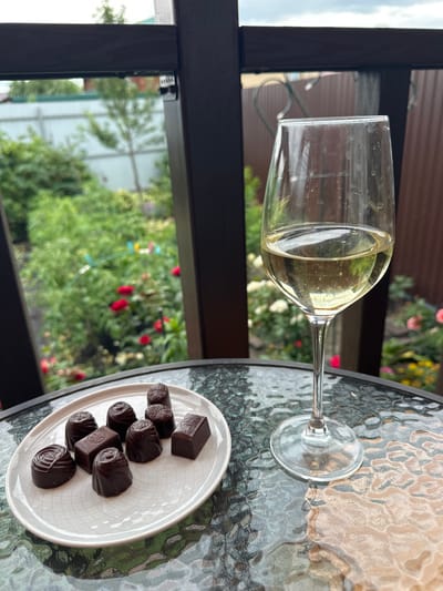 On a textured glass table outdoors, a white plate holds eight dark chocolate confections. To the right of the plate, a wine glass is filled with a pale yellow liquid, likely white wine. The background reveals a garden with lush green foliage and red and white flowers, bordered by a brown wooden fence and a light blue fence. The scene appears to be on a balcony or patio, with a dark wooden railing in the foreground. The location is identified as Zubovo, Russia.