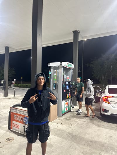 This nighttime scene shows a group of three individuals at a 7-Eleven gas station. A white Honda Civic is parked next to a gas pump, with the driver's side door open. One person, wearing a gray hoodie, is standing next to the car, while another in a green shirt is closer to the gas pump. The third person is near a display stand promoting "on!" nicotine pouches, holding his hands up, and his face is towards the camera. The environment is illuminated by the station's lighting. The location is Homosassa Springs, United States of America, based on the presence of a 7-Eleven gas station.