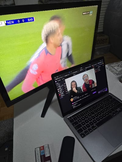 An indoor scene in Krems an der Donau, Austria, depicts a leisure setup with multiple screens. A large, black-framed television displays a soccer match, showing the score "NEW 0 0 BAR" and a game time of "47." The channel logo "ROCKSPORT LIVE" is visible in the top right. A player in a pink jersey with light, short hair is prominently featured on the screen, partially obscured by a blurry player in a striped jersey behind him, against a green field background. Below and to the right of the television, a silver MacBook Pro laptop is open, displaying a Twitch stream or video conference featuring two males: one with dark hair wearing a dark top on the left, and another with glasses wearing a red and black patterned top on the right. The laptop rests on a light gray tabletop. To the left of the laptop, a black, elongated remote control is present. Further left on the table is a rectangular packet or box, possibly a tobacco product, displaying a health warning in a European language, which appears to be "RAUCHEN IST TÖDLICH" (Smoking is deadly). In the background to the right, a dark, upholstered object (likely a couch or chair) is visible, along with a light-colored box labeled "BOOST" and a clear glass object, possibly an ashtray. The environment suggests a living area or personal entertainment space.