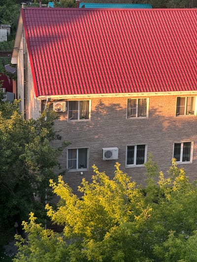 The image shows the facade of a brick building with a red tiled roof, illuminated by the warm glow of the setting sun. The building has multiple windows, some with curtains, and at least two outdoor air conditioning units are visible. Lush green trees, partially illuminated by the sunlight, fill the foreground and partially obscure the lower floors of the building. In the background, glimpses of other buildings with different colored roofs and fencing can be seen, suggesting a residential area. The scene is likely captured from an elevated perspective, looking down onto the building and surrounding foliage. The presence of air conditioning units and the architectural style suggest a temperate climate. Based on the provided context, this scene is located in the city of Khabarovsk, Russia.