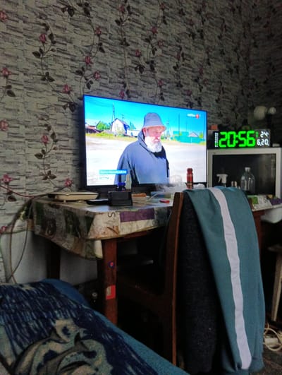 The image depicts an indoor scene, likely a living room or kitchen in Dobryanka, Russia. A television screen displays a news program featuring a man with a beard wearing a hat and a blue hooded jacket. Text on the screen indicates it is "РОССИЯ 1" and "ВЕСТИ." To the right of the television, a digital clock shows the time as 20:56. The room has patterned wallpaper with floral motifs. A wooden table is covered with a patterned tablecloth and cluttered with various items, including a remote control, a bottle of liquid, and what appears to be a router or modem. A chair with a teal and white striped blanket draped over it is positioned in the foreground. Part of a bed with a blue patterned blanket is visible in the lower left corner. The overall impression is of a lived-in, domestic space.