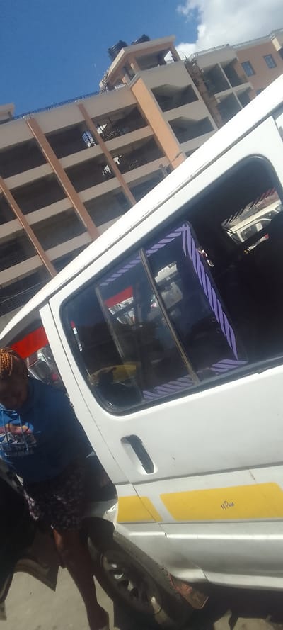The scene depicts an urban environment in Naivasha, Kenya, featuring a white passenger van and a multi-story building under a clear blue sky with scattered clouds.

In the foreground, a white van, consistent with a Kenyan matatu, is oriented diagonally, occupying the lower half of the frame. The vehicle's right side is visible, displaying two horizontal yellow stripes on its lower body panel, with faint handwriting "14 Pass" discernible above the rear yellow stripe. The windows are dark-tinted, and the rear passenger window frame features a vertical purple decal with a diagonal striped pattern. A black exterior door handle is present on the sliding door. The front right tire, with a silver hubcap, is partially visible at the bottom of the image.

An individual is positioned next to the partially open sliding door of the van. This person has reddish-brown braided hair and is wearing a blue long-sleeved hooded sweatshirt with partially visible white text, likely "OLD NAVY CALIFORNIA." They are also wearing patterned shorts. The individual is leaning downwards and inwards towards the van's interior.

The background features a multi-story building, appearing to be a concrete structure with beige or light brown vertical pillars and white horizontal bands or balustrades. Multiple open levels or balconies are visible. On the building's flat rooftop, two black cylindrical water tanks are mounted on an elevated platform, and a thin antenna or pole extends upwards. The overall impression is an active urban setting with ongoing daily activities.