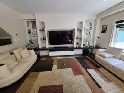 This is an image of a well-lit living room in a modern home, likely in Kitchener, Canada. The scene is centered around a large flat-screen television mounted on the wall, flanked by decorative niches filled with various figurines and artwork. A dark entertainment console sits below the TV.
Two large, cream-colored sectional sofas are positioned in the room, one on the left and one on the right, facing the television. They are adorned with plush throw pillows. A large, abstract patterned rug with geometric shapes in shades of brown, beige, black, and red covers a significant portion of the dark hardwood floor. A small, bright yellow ball rests on the rug in front of the television.
To the left of the TV, a tall, abstract piece of artwork hangs on the wall. To the right, a small framed picture of a historical figure in a red and gold uniform is displayed near a window with blinds. A decorative hookah is also visible on a surface to the right of the entertainment console, suggesting a potential cultural element. The overall atmosphere is one of a comfortable and stylish living space, likely captured during the daytime given the natural light streaming in. There is no visible text in the image.