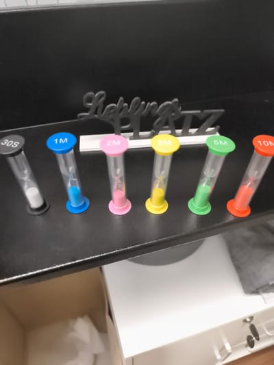 The image displays a collection of six hourglass timers arranged on a black shelf. Each hourglass is a different color and labeled with a time duration: 30S, 1M, 2M, 3M, 5M, and 10M. The sand inside each hourglass is in various stages of falling, indicating they are in use or have been recently used. Behind the hourglasses, a black decorative word art piece is visible, with the text "Lieblingszeit" and "A-Z" clearly legible. The overall scene is indoors, with a hint of a cardboard box and white furniture in the background, suggesting a domestic or office setting. The lighting appears to be artificial and even. There are no people visible in the image. The context provided suggests the location is Troisdorf, Germany.