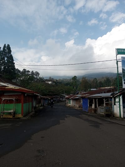 The image depicts a street in a rural or mountainous area. A paved road runs through the center of the frame, flanked on both sides by small, weathered buildings or shops. The buildings appear to be simple structures, some with corrugated metal walls and rudimentary stalls or counters. The sky is partly cloudy with a mix of blue sky and heavy cloud cover, suggesting either early morning or late afternoon light. In the background, a forested hill or mountain range is visible, partially obscured by clouds or mist. There is a motorbike on the road traveling away from the camera. Vegetation is sparse. The location appears to be a remote or economically challenged area. There is no visible indication it is specifically the location of Mya Sit Kaing Ywar Thit, Myanmar. There is a street sign with an indecipherable word or words on it.