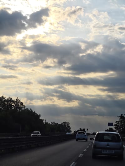 The image depicts a highway in Acerra, Italy, with a dramatic sky filled with sun rays piercing through dense clouds. Several cars are visible on the road, moving in the same direction. In the foreground, a silver hatchback is in the right lane, its license plate reading "KDA 08204". To its left, another white hatchback is in the left lane. Further ahead, more vehicles can be seen on the highway, including a white SUV in the distance. The sides of the highway are lined with trees, creating a dark, silhouetted barrier against the bright sky. Traffic signs and lights are also present along the roadside. The overall scene suggests a journey during a cloudy day with intermittent sunlight.