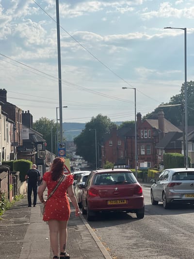 An image of a street scene shows a red-haired woman in an orange dress standing on the sidewalk next to parked cars. Behind her are buildings and a street sign showing a speed limit of 30. The sky is light blue with clouds. In the background, there are distant hills. A man in dark clothing is walking away from the camera, closer to the building. Streetlights line the road. The scene appears to be a typical residential street on a sunny day.
