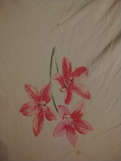 This is a close-up, static, medium shot image of three pink flowers with green stems painted on a light-colored fabric. The fabric appears to be textured, possibly linen or cotton, and shows some wrinkling. The painting style is simple and somewhat abstract, with visible brushstrokes. The flowers have five petals each and appear to be in various stages of bloom. The lighting is diffused, and there are no shadows visible. There is no discernible location in Bamberg, Germany, as the image only depicts the painted fabric.