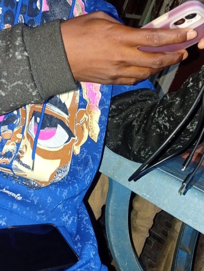 A close-up shot shows a person's hands and arm resting on a blue surface, with a phone being held in the right hand. The person is wearing a blue hoodie with a graphic design of an anime-style eye and hair. A black fabric is visible on the left side of the frame, and several black cables are arranged on the blue surface. The background is blurred, suggesting an indoor setting. The image does not contain any explicit indicators of location within Ukpor, Nigeria.