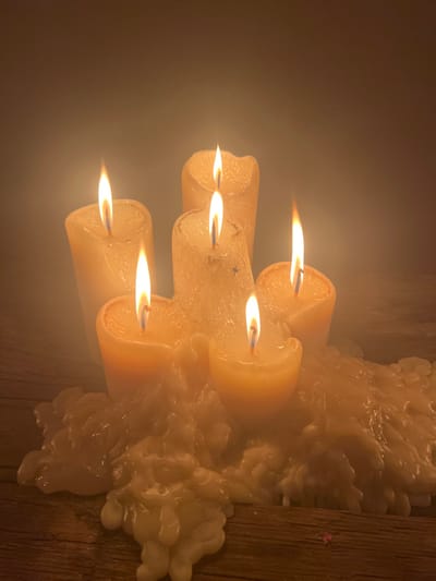 A collection of six lit candles are grouped together, displaying varying burn lengths. The candles are a creamy, off-white color, with visible signs of dripping wax that have accumulated around the base, creating a congealed mass. The bright flames of the candles contrast with the darker background, and light reflects off the wax surfaces, giving a soft glow. The arrangement is attached to a rough-hewn wooden surface.