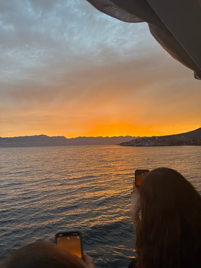 The image shows a sunset over water, with a rocky coastline visible in the middle ground. The sky transitions from a cloudy white to a vivid orange hue as it approaches the horizon. Two individuals are partially visible on the right side of the frame, each holding a smartphone up as if taking a picture of the scene. A curtain is visible in the upper left corner. The location is near water and coastline in Unknown, Mexico.