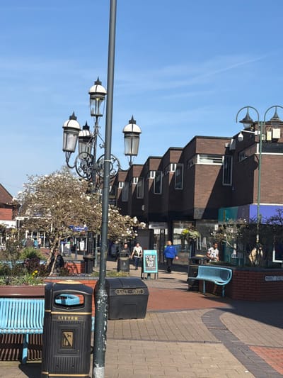 This image captures a bright, sunny day in a pedestrianized public area in Crewe, United Kingdom, specifically near Market Street, indicated by a street sign visible on the right.
The foreground is dominated by a tall, dark grey lamppost with an ornate four-lantern fixture; each lantern features a white dome and dark metal framework, with one lantern exhibiting a cracked glass panel. To the left of the lamppost, a light blue slatted metal bench is positioned next to a black "LITTER" bin displaying a dog waste symbol. To the right of the lamppost base, a black "SALT - GRIT" bin is present. The ground is paved with multi-toned block pavers in grey, brown, and reddish-brown patterns.
In the midground, two trees with white blossoms are situated within low brick planters. Beyond these, a pedestrian walkway extends into the background, flanked on the right by a continuous structure of brown brick commercial buildings. These buildings feature multiple levels, characterized by sloped sections and regularly spaced vertical, angled windows. Shopfronts are visible at ground level, with one featuring a partial "High Street" sign and another "TO LET" sign.
Several individuals are present on the walkway. A person wearing a blue top and dark trousers walks away from the viewer, centrally located. Behind them, another individual in a white top with dark trim walks towards the camera. Further right, a person in a patterned short-sleeved shirt is seated on a light blue slatted metal bench situated on a raised brick planter. An A-board sign displaying "50GB SIM £13" stands near the walkway. Another street light with two arched lamps is visible on the far right. The sky above is clear and blue.