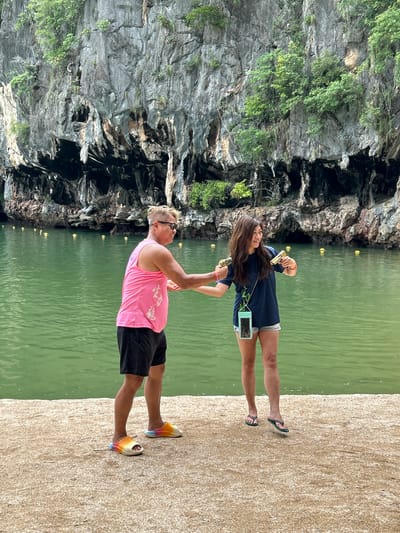 In Krasom, Thailand, a man and a woman stand on a sandy shore next to a body of green water. The man, wearing a pink tank top and black shorts, offers his hand to the woman, who is dressed in a navy blue t-shirt and denim shorts. She holds a gold toy gun in each hand. Behind them, a towering limestone cliff face is covered with lush green vegetation and features several dark cave openings. Yellow buoys float on the water's surface, marking the edge of the swimming area.