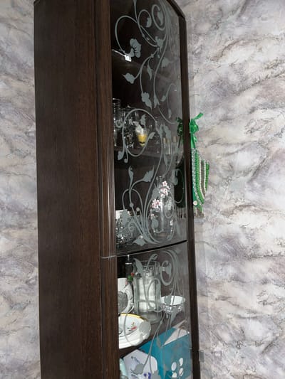 A tall, dark wood cabinet with glass doors sits against a marble-patterned wall in Abzakovo, Russia. The glass doors are etched with a floral design. The cabinet contains various glassware, including champagne flutes and decorative vases, some with flowers. A string of green and white beaded jewelry hangs from a ribbon on the right side of the cabinet. Below the glassware, several white teacups with floral patterns and gold rims are visible, along with a blue box. The lighting is dim, casting shadows on the cabinet and wall.