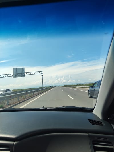 The image shows a view from inside a vehicle on a highway. The sky is bright blue with scattered white clouds. The highway has multiple lanes and a metal guardrail runs alongside it. There are several cars visible on the highway. In the foreground, the dashboard of the vehicle is visible. There is a large overhead sign structure on the left side of the highway. The landscape appears to be somewhat flat with distant hills. The scene is consistent with driving on a highway outside of a city, and the visual cues do not specifically place it within Lusakert, Armenia.