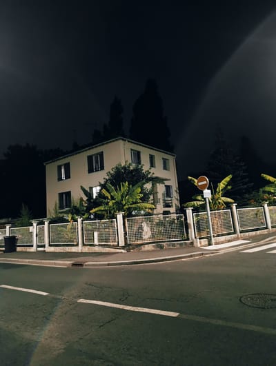 The image captures a scene at night on a street corner in Villeneuve-Saint-Georges, France. In the background, a two-story building with illuminated windows stands behind a white metal fence. Large banana plants are visible in front of the building, adding a tropical element to the urban landscape. A "No Entry" sign is prominently displayed on a pole near the fence. The foreground shows a dark asphalt road with white lane markings and a pedestrian crossing. A faint rainbow-like refraction is visible on the road surface, possibly from a light source. A black trash can is situated near the fence on the left side of the image. The overall atmosphere is quiet and illuminated by artificial lights, typical of a residential street at night.