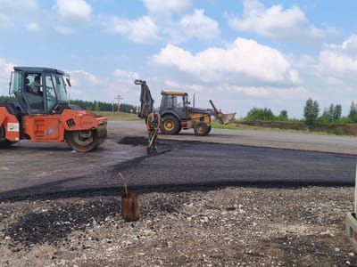 The image shows a road construction scene on a sunny day. In the foreground, a roller is compacting freshly laid asphalt. A worker in a camouflage uniform and a high-visibility vest is using a shovel to spread the asphalt. Behind them, a yellow backhoe is parked, with its bucket raised. The background features a road, trees, and a cloudy blue sky. In the distance, there appears to be a wooden cross on a small hill. This scene is located in Borskoe, Russia.