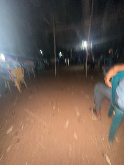 A blurred image shows an outdoor scene at night. People are present, with one person in the foreground wearing a teal shirt and gray pants. The ground is dirt, scattered with what appears to be debris. In the background, there are rows of seats and poles with lights, suggesting an event or gathering. A yellow chair is visible in the lower portion of the frame.