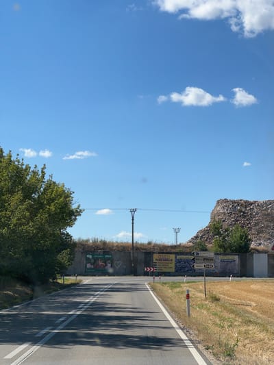 The image depicts a rural roadside scene under a clear blue sky with scattered white clouds. A paved road with double white lines curves to the left. On the left side of the road, a large, leafy green tree casts shadows on the pavement. On the right side of the road, dry, yellowed grass borders the asphalt, and a pile of rubble or debris rises in the background, forming a large mound. A concrete wall runs along the right side of the road, adorned with several billboards and signs. One billboard features a graphic of produce and text that appears to be an advertisement. Another sign points to "NETAGAS s.r.o." and "FRUJO" with distances indicated. A white heart shape is drawn on the concrete wall. Utility poles with wires are visible in the distance, and a large pile of construction debris or waste material is visible on the right side of the frame, dominating the background. The scene is set in Tvrdonice, Czechia, as indicated by the visible signage.