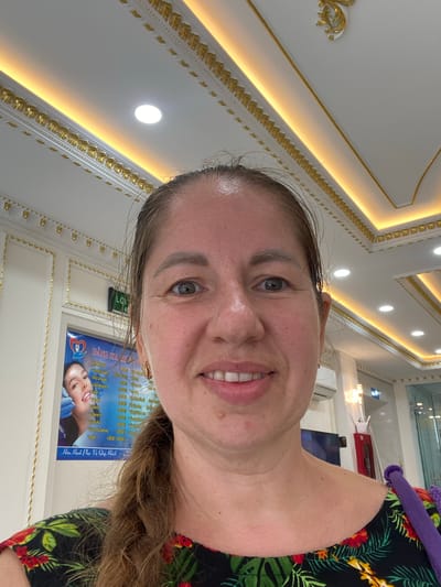 A woman with blonde hair and fair skin, wearing a black shirt with a floral pattern, takes a selfie in what appears to be a dental clinic or beauty salon. The background features a highly decorated white ceiling with gold trim and recessed lighting. A poster on the wall behind her shows a woman's face with a bright smile and a price list in Vietnamese. The overall impression is one of a brightly lit, well-maintained indoor space. The location is identified as Phú Thủy, Vietnam.