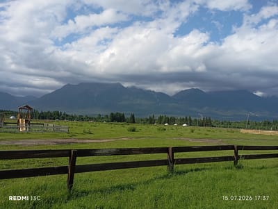 In Khurai-Khobok, Russia, on July 15, 2026, at 17:37, a rural landscape is depicted under a dramatic sky. A dark wooden fence dominates the foreground, stretching horizontally across the frame. Beyond the fence, a lush green meadow unfolds, dotted with scattered trees and several yurt-like structures in the middle distance. A children's playground with a wooden structure and a white picket fence is visible to the left. In the background, imposing mountains rise, their peaks partially obscured by heavy, dark clouds that contrast with patches of blue sky. The overall scene suggests a remote, natural setting with evidence of habitation.