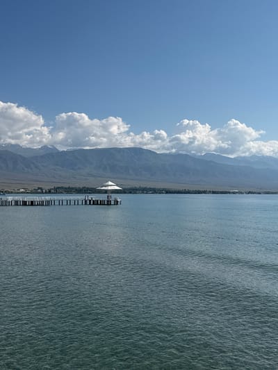 The image captures a serene view of a pier extending into a tranquil body of water, with a majestic mountain range forming a distant backdrop. The pier, constructed from white planks and supported by numerous vertical posts, juts out into the calm, blue water. At the end of the pier, a white, multi-paneled umbrella provides shade for a small group of people, some of whom are standing or sitting. A few more figures are visible along the length of the pier. In the foreground, the water exhibits a gentle ripple pattern, and a series of small, white buoys are strung across the surface, marking a boundary or area. The mountains in the distance are layered with varying shades of blue and brown, their peaks softened by the atmospheric haze. The sky above is a clear, bright blue, punctuated by fluffy white clouds scattered across the upper portion of the frame. The scene suggests a peaceful, recreational setting, likely a resort area on the southern shore of Lake Issyk-Kul, near the village of Koshkol', Kyrgyzstan.