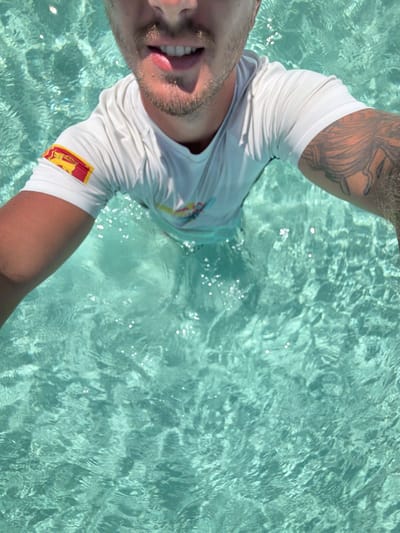A male subject is depicted partially submerged in clear, turquoise water in Gulhi, Maldives. His face, from the nose down, is visible, showing light facial hair and a slight smile with teeth exposed. He wears a wet white short-sleeved top. A rectangular patch on the left sleeve displays a red background with a yellow lion holding a sword. His right arm features a dark, intricate tattoo with organic, tentacle-like patterns. The water's surface is disturbed by ripples and reflects sunlight. The subject's outstretched arms indicate he is holding a device to capture a self-portrait.