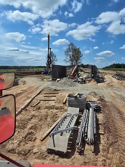 The image depicts a construction site in Piecnik, Poland, under a bright, partly cloudy sky. A large drilling rig stands prominently in the background, suggesting deep foundation work or soil exploration. Adjacent to it, a red crane is visible, hinting at ongoing heavy lifting operations. The ground is uneven and consists of disturbed earth and piles of sand and gravel, typical of an active construction zone. Several stacks of metal scaffolding and construction materials, including pipes and structural elements, are organized in the foreground, indicating preparation for or progress in building. The scene appears to be a large-scale infrastructure project, possibly for a bridge or a significant building, given the scale of the equipment and materials. The overall impression is one of active construction and industrial activity in a semi-rural or suburban setting.