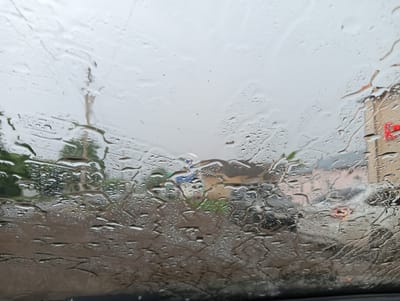The image shows a view through a wet car windshield. Water droplets and streaks distort the scene outside, which appears to be an urban environment under overcast skies. The background shows blurred buildings, some with windows and architectural details, and hints of greenery. A red traffic signal is visible on the right side. The foreground is dominated by the texture of the raindrops on the glass. The specific location in Nizhnetroitsky, Russia, cannot be determined solely from this image.
