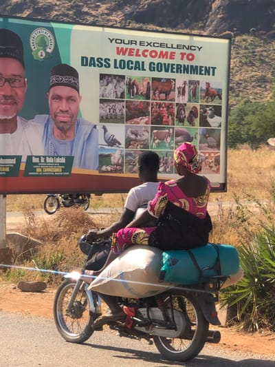 This image captures a daytime scene on a paved road, in or near Bundot, Nigeria, with mountainous terrain in the background. A large billboard dominates the upper portion of the frame, displaying "YOUR EXCELLENCY WELCOME TO DASS LOCAL GOVERNMENT" and identifying "BAUCHI STATE OF NIGERIA." The billboard features portraits of two men, identified as Governor Alh. Abibukar S. Mohammed and Hon. Dr. Bala Lukshi, Commissioner for the Ministry of Livestock, Bauchi State. The right side of the billboard contains a grid of photographs depicting various livestock and poultry, including cattle, goats, horses, chickens, pigeons, and turkeys.
In the mid-ground, two individuals are riding a motorcycle from left to right. The driver is a male with dark skin, wearing a white t-shirt. Behind him, a female passenger with dark skin is seated, wearing a vibrant pink, yellow, and black patterned dress and a matching headwrap. She carries a dark bag. The motorcycle is heavily loaded: a large, light-colored sack is strapped to the rear seat beneath the female passenger, and a dark teal cylindrical object is secured horizontally at the very back. Another motorcycle is partially visible in the middle distance, positioned below the billboard, appearing to be stationary or traveling in the opposite direction. Dry vegetation and sparse greenery line the roadside.